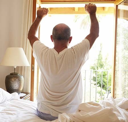 Man waking up in the morning who is sitting in bed & stretching his back.