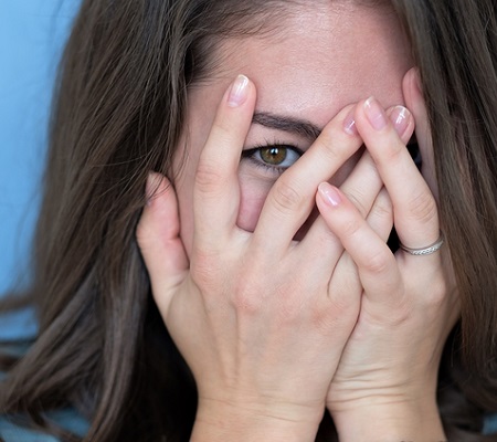 Studio shot of brunette girl hiding eyes under hand while feeling ashamed. Caucasian young woman in white shirt covering face with hand. Human facial expressions and emotions
