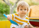 kid in yellow striped shirt with blue scoop shovel crouching in a sandbox