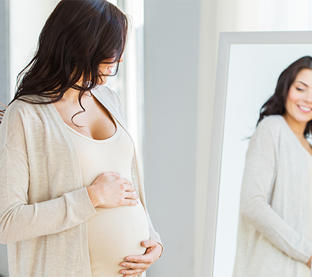A pregnant women holding her belly and looking in the mirror with a smile.